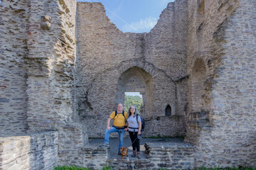 Obraz na płótnie Canvas Mature tourist couple smiling and looking at the camera next to their brown dog, Brandenburg castle in ruin with stone walls, window in the background, sunny summer day in Luxembourg