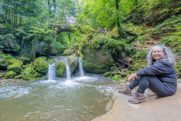 Female hiker tourist sitting on rock, Schiessentümpel waterfall on Black Ernz river and abundant green wild vegetation in background, greyish hair, looking at camera, Mullerthal Trail, Luxembourg - obrazy, fototapety, plakaty
