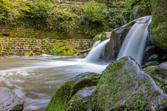 Side view of the Schiessentümpel waterfall with water flowing between the rocks in the Black Ernz river, brick wall with moss in the background, Mullerthal Trail, Luxembourg. Long exposure image
