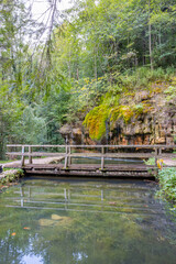 Fototapeta na wymiar Wooden bridge over a spring with crystalline calcareous water, mossy sandstone rock formation with a waterfall in the background, Kallektuffquell travertine source, Mullerthal Trail, Luxembourg