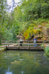 Mullerthal Trail, tourist couple standing on a wooden bridge over the basin, Kallektuffquell waterfall, water flowing between rock formations with green moss in the background, sunny day in Luxembourg