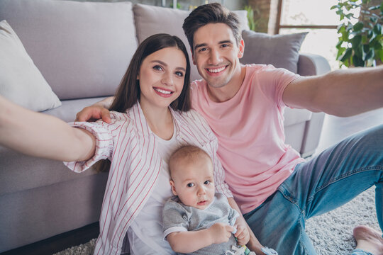 Self-portrait of attractive cheerful full family handling growth baby sitting on carpet embracing cuddling at home indoors