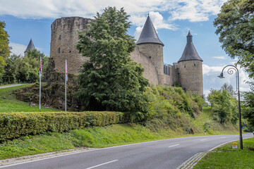 Fototapeta na wymiar Asphalt country road next to the medieval castle of Bourscheid on a rocky hill, a tree, green grass and flagpoles with two flags, stone walls, watchtowers, sunny day with a clear blue sky, Luxembourg