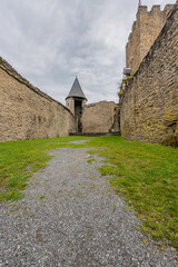 Fototapeta na wymiar Dirt path in the direction of the watchtower along the green grass, deteriorated stone walls in the medieval castle of Bourscheid, cloudy day with a sky with gray clouds, Luxembourg