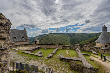 Outdoor ruin at the medieval castle of Bourscheid, ruined stone walls, towers, roofs and the valley with trees and a cloud covered sky in the background, seen from the uppermost courtyard, Luxembourg