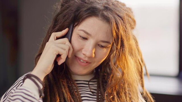 Tilt up of smiling young woman in striped sweater with dreadlocks answering call on cell phone while working or learning online using laptop at home. Remote work at quarantine concept