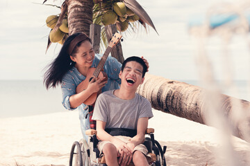 Young man with disability and mother smiling and singing, playing music therapy on the beach,...
