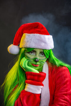 woman disguised as christmas grinch, sitting in studio black background with santa claus costume green wig lady grinch with smoke and christmas lights