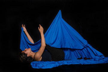 Latin woman with indigenous characteristics with her body wrapped in a blue cloth lying down raising arms on a black background
