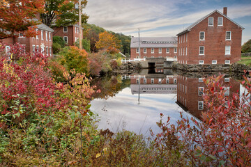 Fototapeta na wymiar Colorful autumn scene at historic 19th-century textile mill town in Monadnock region of New Hampshire. Picturesque village of Harrisville with reflection of sky and red brick buildings on water.