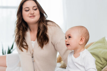 Cheerful baby girl sitting on bed near blurred mom at home.