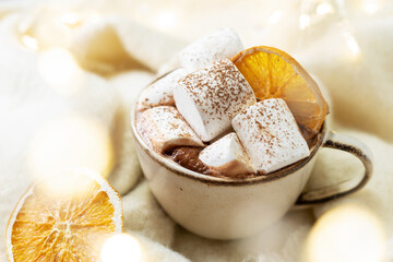 Traditional Christmas cocoa in a ceramic cup with marshmallows and orange chips on a light background with bokeh