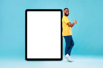 Positive black guy showing thumb up gesture next to huge digital tablet on blue studio background, mockup