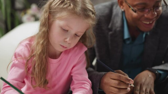 Concentrated little girl draws picture with pencil at homeschooling with African-American native speaker private teacher at home closeup slow motion