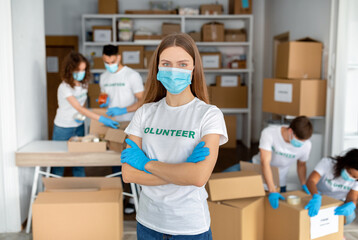 Fototapeta na wymiar Volunteer organization. Young woman in medical mask and gloves posing with folded arms while working at charity center