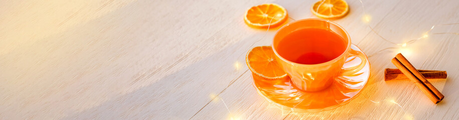 Orange cup of tea, sinnamon, slice of dried orange, lights on wooden background, banner, copy space