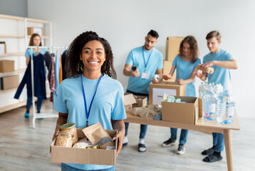 Happy african american female volunteer in uniform holding cardboard box with food donation, working in charity center