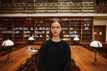 Portrait of a beautiful woman in dark clothes standing in an atmospheric library and looking at the camera with a serious face on the background of bookshelves.