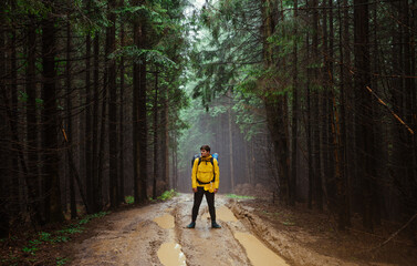 Obraz premium Male hiker stands on a winding road in the mountains with a backpack on his back against the backdrop of a beautiful misty forest after the rain.