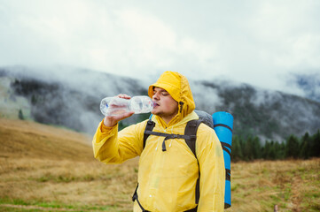 Hiker in a yellow raincoat on a hike in the rain drinks water from a bottle on a background of beautiful mountain overcast views