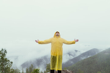 Happy hiker man climbed to the top of the mountain in the rain, raised his hands up and rejoices against the backdrop of a beautiful landscape.