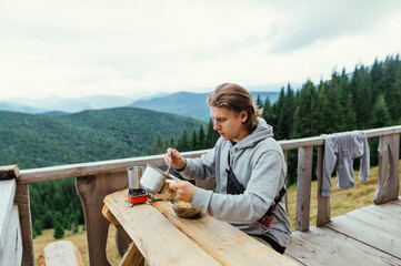 Fototapeta premium Guy tourist lunch on the terrace of a wooden bungalow on a background of mountains. The tourist spent the night in a mountain house, eating pasta for breakfast.