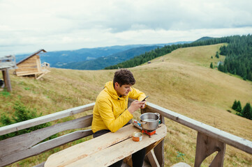 Man prepares food on the terrace of a wooden house in the mountains with a burner and uses a smartphone.