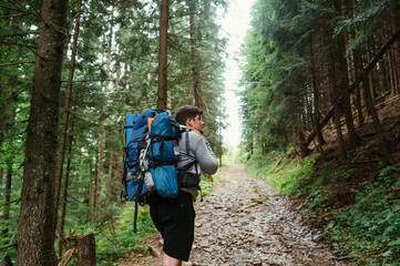 Male tourist with a backpack on his back climbs the mountains on a forest path and looks away.