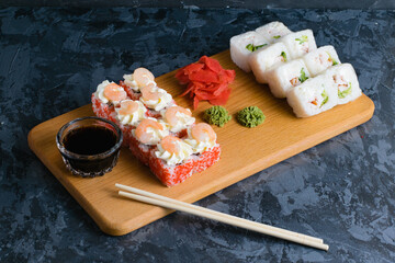 Appetizing and delicious sushi rolls with shrimp, cheese, caviar, chicken, soy sauce, nori, wasabi and ginger on a wooden board on a dark background