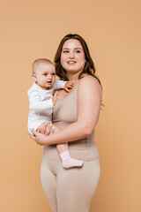 Happy woman with overweight holding baby and looking away isolated on beige.