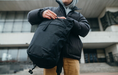 Man in casual clothes opens a black backpack on the street on an architecture background.