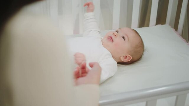 Smiling cute little baby girl very excited hugging hands of her mother while is laying down on the baby cot