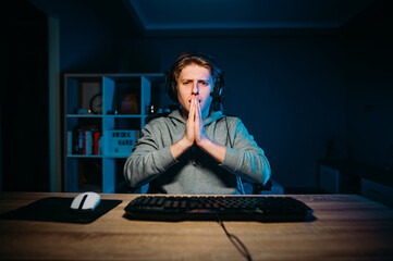 Guy gamer sits in a headset at home at night at the computer and prays for his team looking into the camera with despair on his face. © bodnarphoto