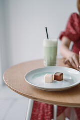 Variety chocolate pralines on white plate and fresh green matcha latte tea in ceramic mug on wooden table at modern style cafe.