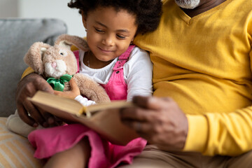 Smiling small african american girl with toy reads book with elderly grandfather on sofa at home interior, cropped
