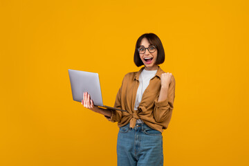 Overjoyed lady raising clenched fist and celebrating success with laptop computer, standing over yellow background