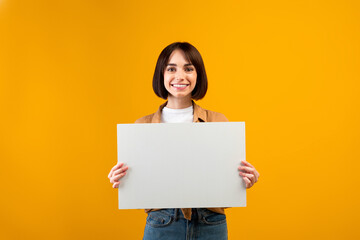 Happy young lady holding white board for advertisement or text, standing over orange studio background, free space