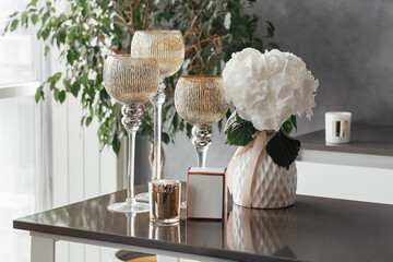 Three glass candlesticks on high legs stand on the table next to a vase with flowers and a gold...