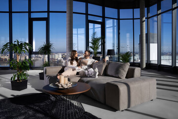 Attractive woman and handsome man relaxing on cozy couch, looking on each other and chatting. Happy couple spending free time together at home. Panoramic windows at apartment.