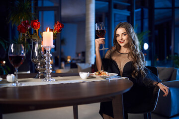Gorgeous caucasian woman in elegant black dress sitting at table with glass of red wine and looking at camera. Romantic mood and atmosphere at home. 