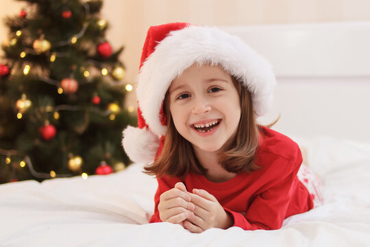 Happy little girl in Santa hat and Christmas pajamas lies on the bed in the bedroom with a Christmas tree