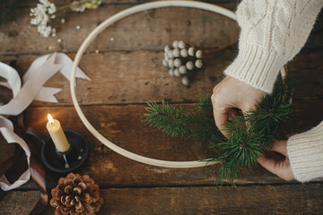 Hands making modern christmas wreath on rustic table with fir branches, brunia herb, ribbons, round wooden hoop, scissors, thread, candle. Atmospheric moody image. Winter holidays preparation