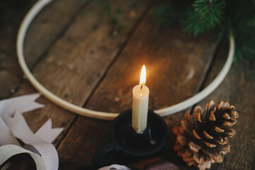 Stylish burning candle and pine cone on background of fir branches, ribbons, round wooden hoop on...