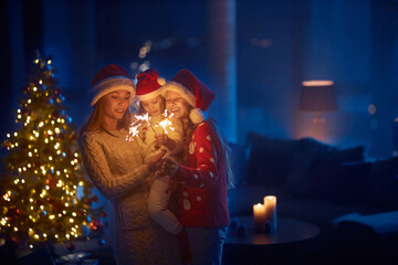 Obraz na płótnie Canvas Happy caring mother and her two little daughters using sparkles during new year celebration. Young woman and girls in santa hats standing at dark living room near shiny christmas tree.