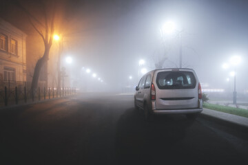 Silver minivan in dense fog on a night street in the city. Soft focus. The light of street lamps along the road in the fog. Car on a foggy autumn night in the city.