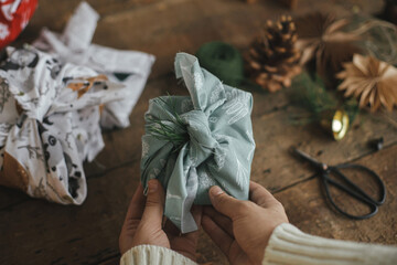 Hands holding christmas gift wrapped in fabric on rustic wooden table with ornaments. Atmospheric moody image, nordic style. Merry Christmas! Furoshiki wrap, zero waste holidays