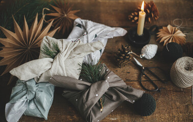 Christmas gifts wrapped in fabric on rustic wooden table with scissors, paper stars, candle,...