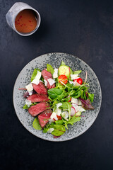 Modern style traditional fried dry aged bison beef rump steak slices with vegetable, lettuce and mustard raspberry dressing served as top view on a Nordic design plate