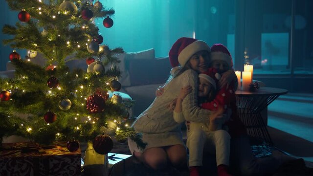 Cheerful mother hugging her little daughters while sitting near beautiful christmas tree with burning garlands. Caucasian family laughing sincerely and clapping in hands. 