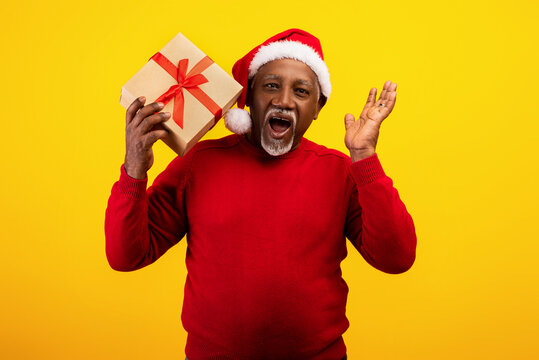 Excited senior black man in red sweater and Santa Claus hat holding gift box, shouting HO HO HO, offering big Xmas sale